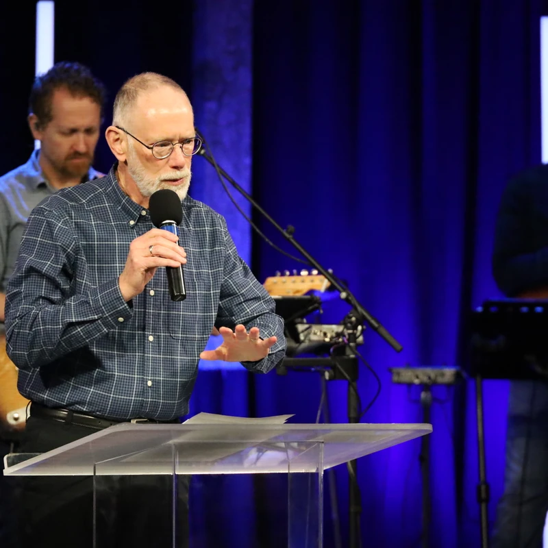 An older man with glasses and a gray beard in a plaid shirt speaks into a microphone while standing at a clear lectern on a stage with a blue curtain.