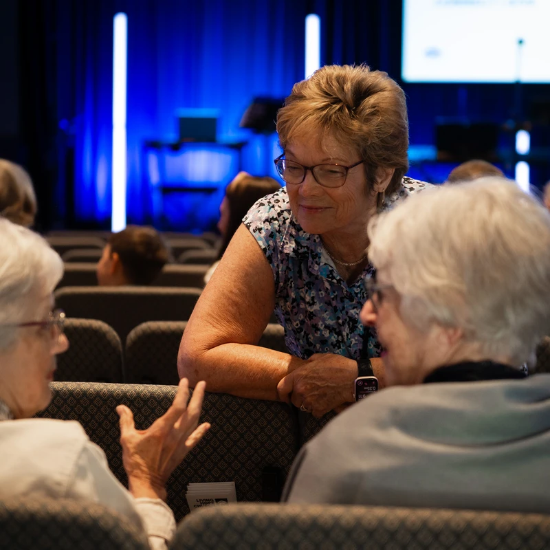 Three older women sitting in auditorium chairs, engaged in a friendly conversation. One woman leans forward to listen as another speaks and gestures.