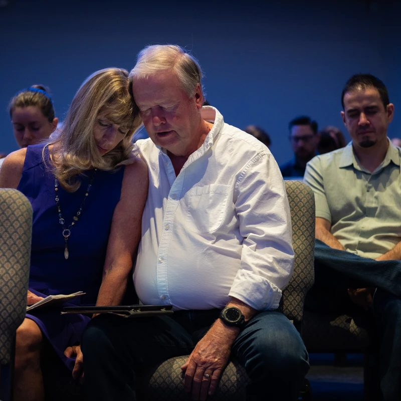 An older couple sits side-by-side in a dimly lit room, their heads touching and eyes closed in a moment of prayer or quiet reflection among other people.