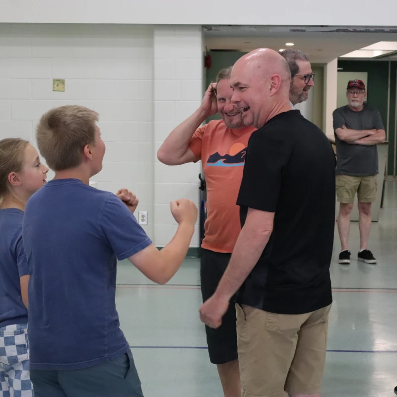 A group of adults and children stand and talk in a community hall. In the foreground, a boy in a blue shirt speaks animatedly to a smiling bald man.