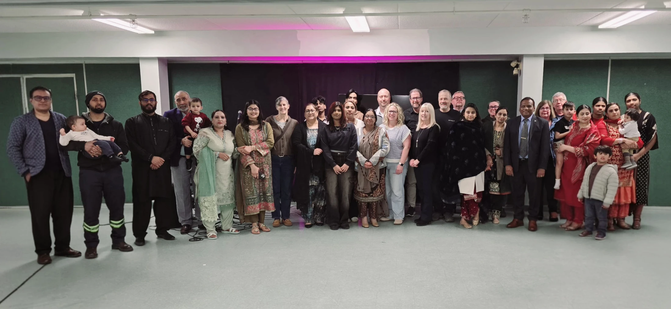 A large, multicultural group of men, women, and children of all ages standing together for a photo indoors in front of a black backdrop with pink lighting.