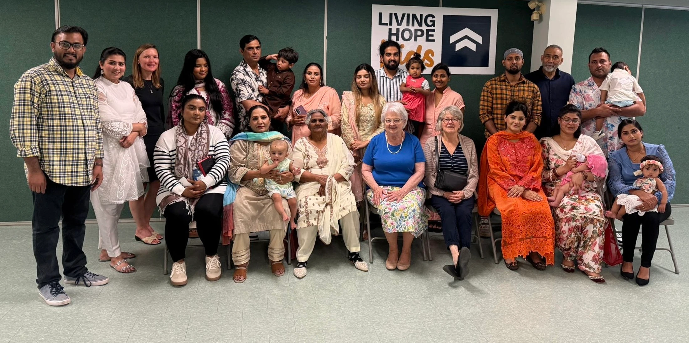 A large, diverse group of adults, toddlers, and babies from the Living Hope community pose for a group photo. Some are seated, and others are standing.