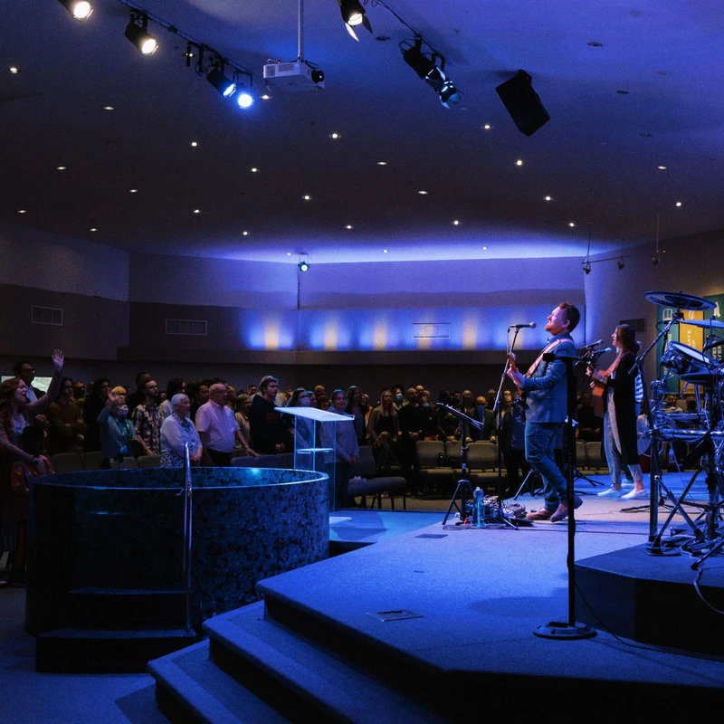 A congregation stands during a worship service, facing a band on a blue-lit stage. A baptismal font is visible next to the stage.