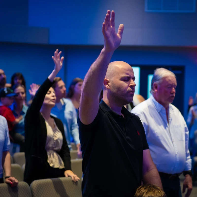 A man with a shaved head and closed eyes raises one hand in worship during a church service. Other members of the congregation are visible behind him.