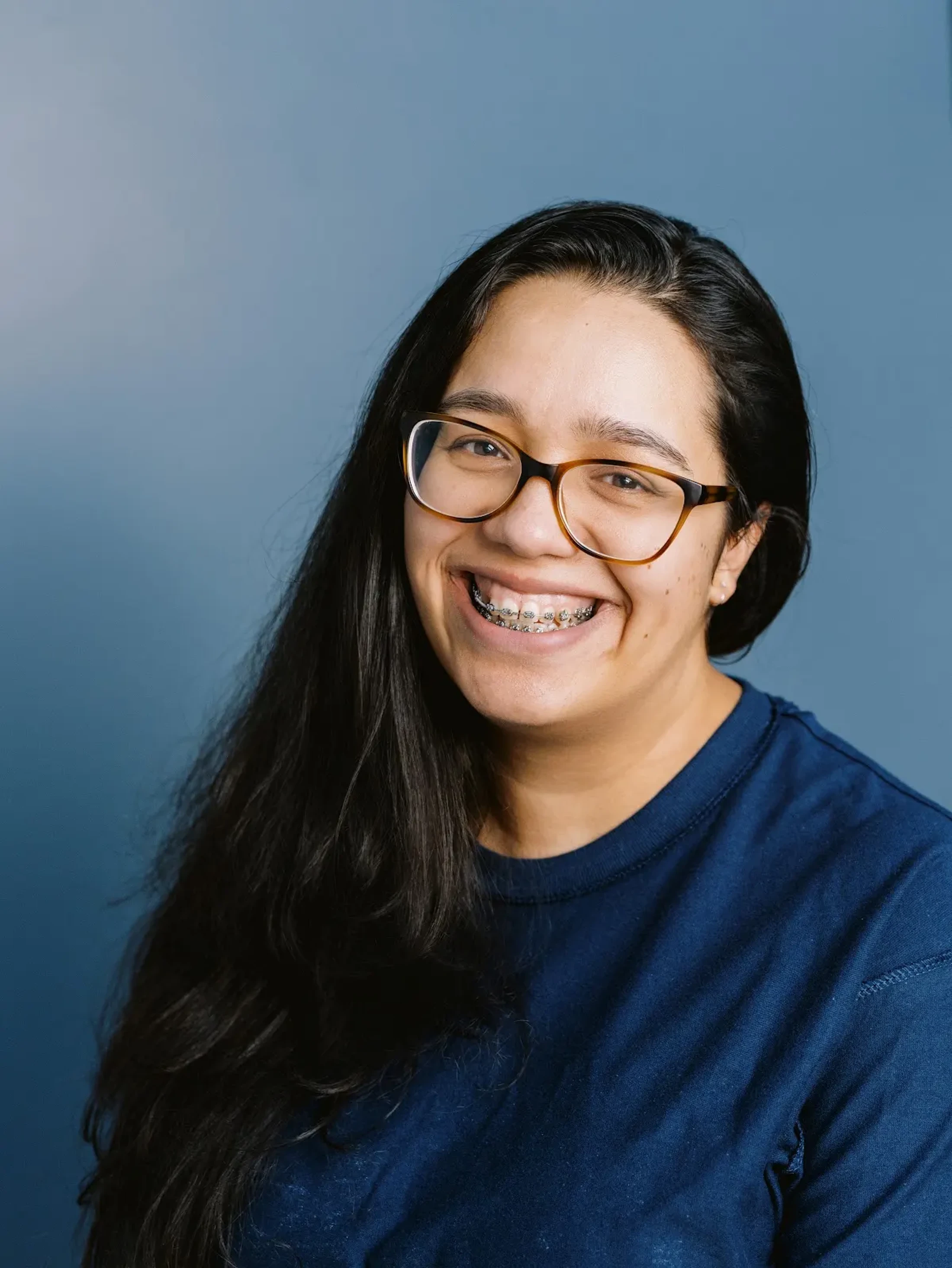 A headshot of a smiling young woman with long dark hair, glasses, and braces, wearing a navy blue shirt against a plain blue background.