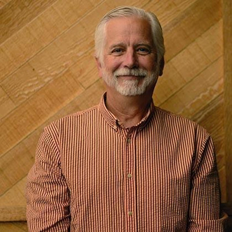 A head-and-shoulders portrait of a smiling older man with white hair and a beard, wearing a striped shirt, standing against a diagonal wood-paneled wall.