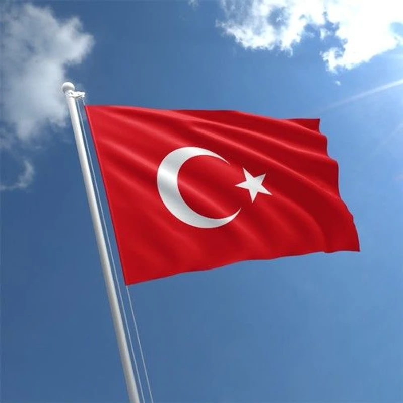 The red flag of Turkey, featuring a white crescent moon and star, is attached to a white flagpole and waving against a bright blue sky with clouds.