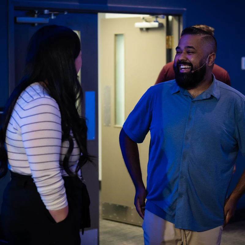 A smiling man with a beard and a small microphone headset talks to a woman with long dark hair in a room with dark blue lighting.