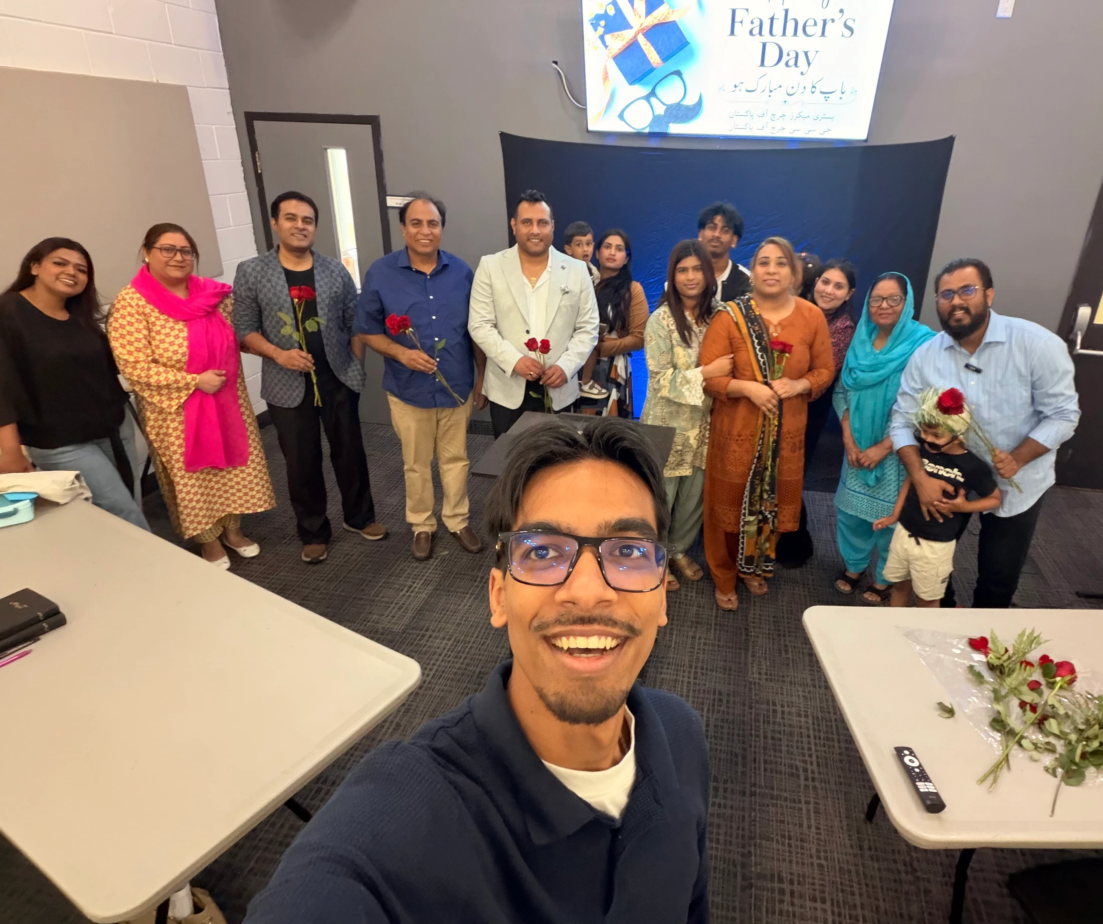 A young man with glasses takes a selfie with a large, happy group of people standing behind him. A screen in the background says 'Father's Day'.
