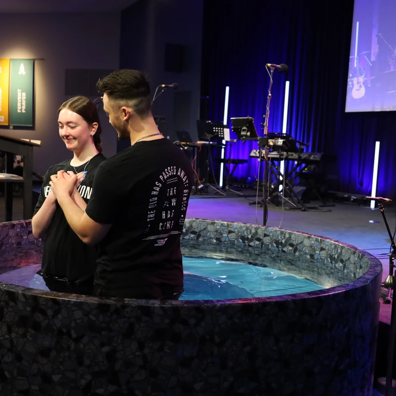 A smiling young woman and a man stand in a baptismal pool on a church stage, holding hands and preparing for her baptism during a service.