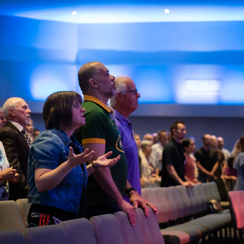 A diverse group of people stand in a modern church with blue lighting. They look upwards, singing and praying during a service.