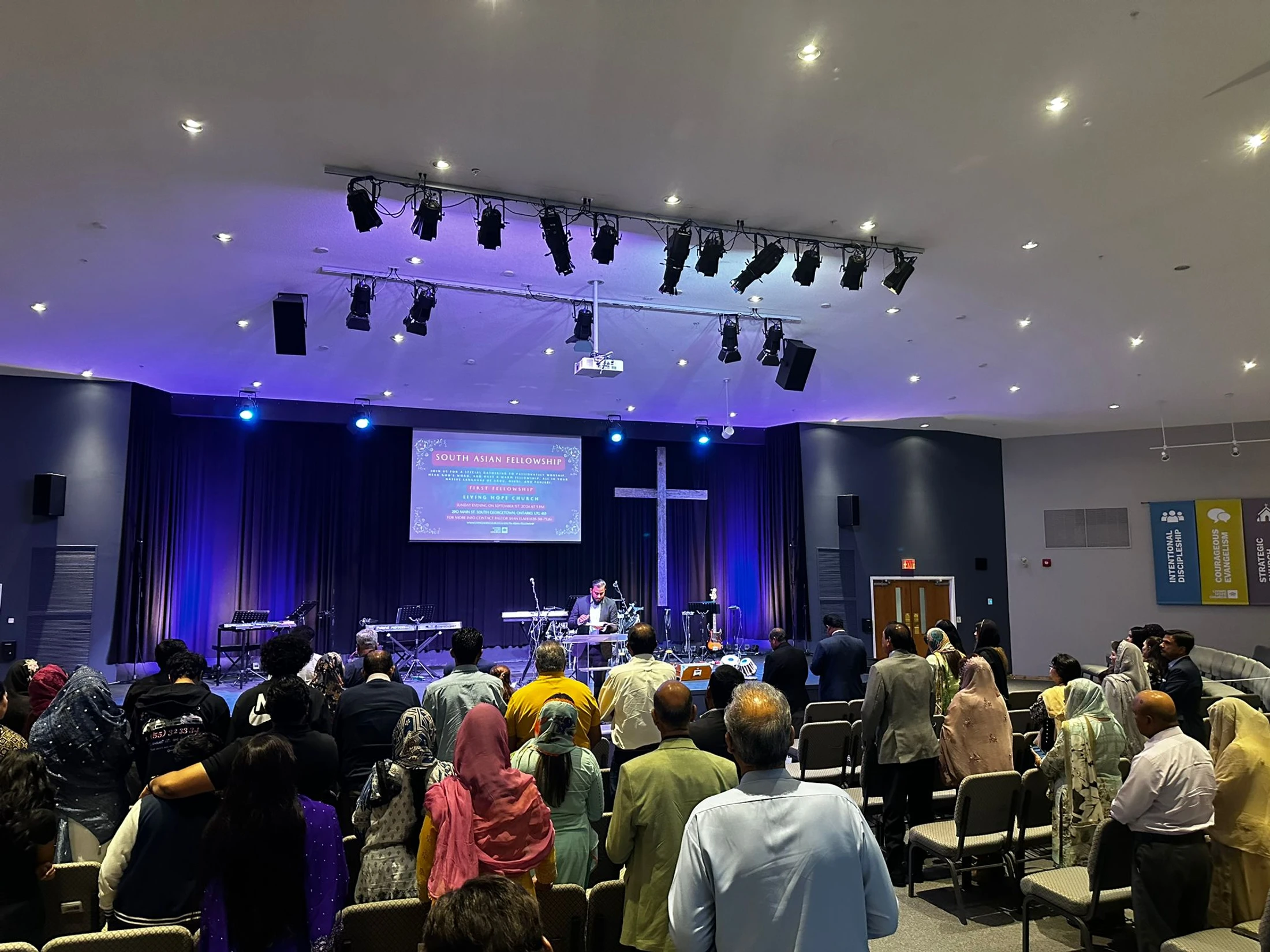 A congregation stands during a South Asian Christian fellowship service, facing a speaker on a stage with a cross and musical instruments.