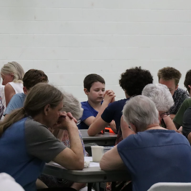 People of all ages sit together at long tables, their hands clasped and eyes closed in a moment of communal prayer.