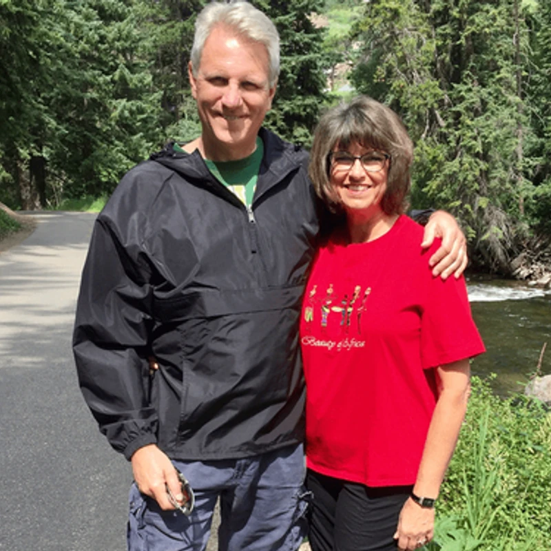A smiling man with his arm around a smiling woman, both standing on a paved path in a forest next to a river.