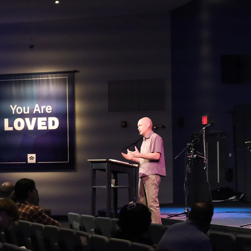 A bald man speaks from a lectern on a stage to a seated audience. Behind him, a large banner with blue and purple lighting reads 'You Are LOVED'.