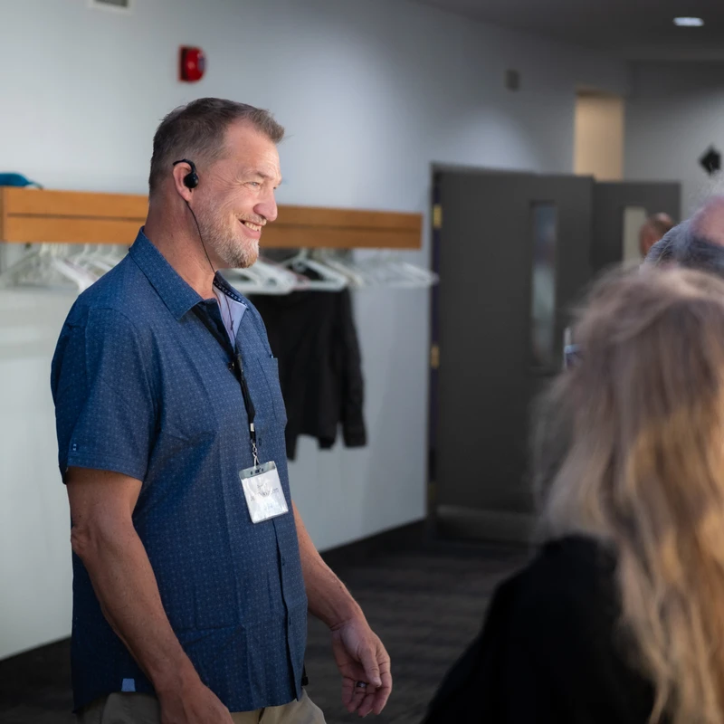A man in a blue button-down shirt with a lanyard and earpiece smiles while interacting with others in a modern building lobby.