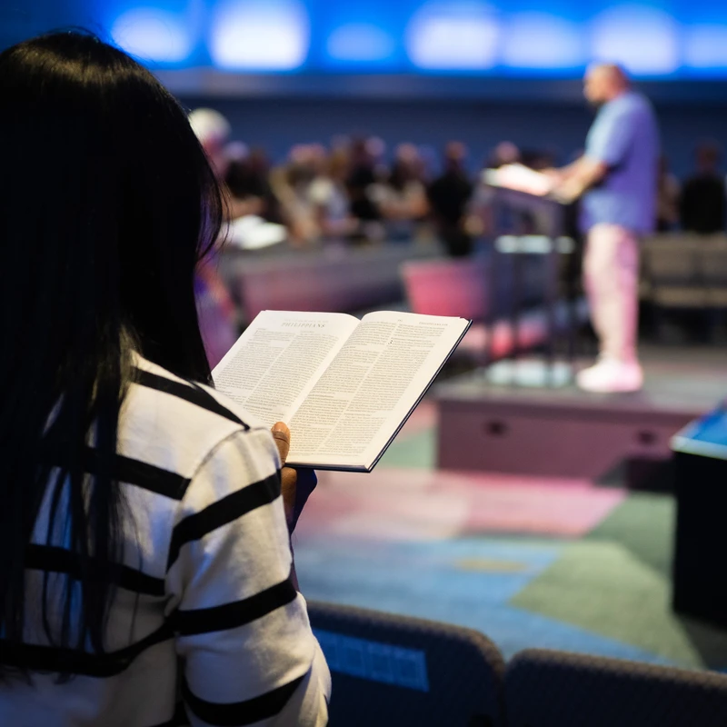A woman with long dark hair, seen from behind, holds an open Bible while listening to a sermon. A pastor and congregation are blurred in the background.
