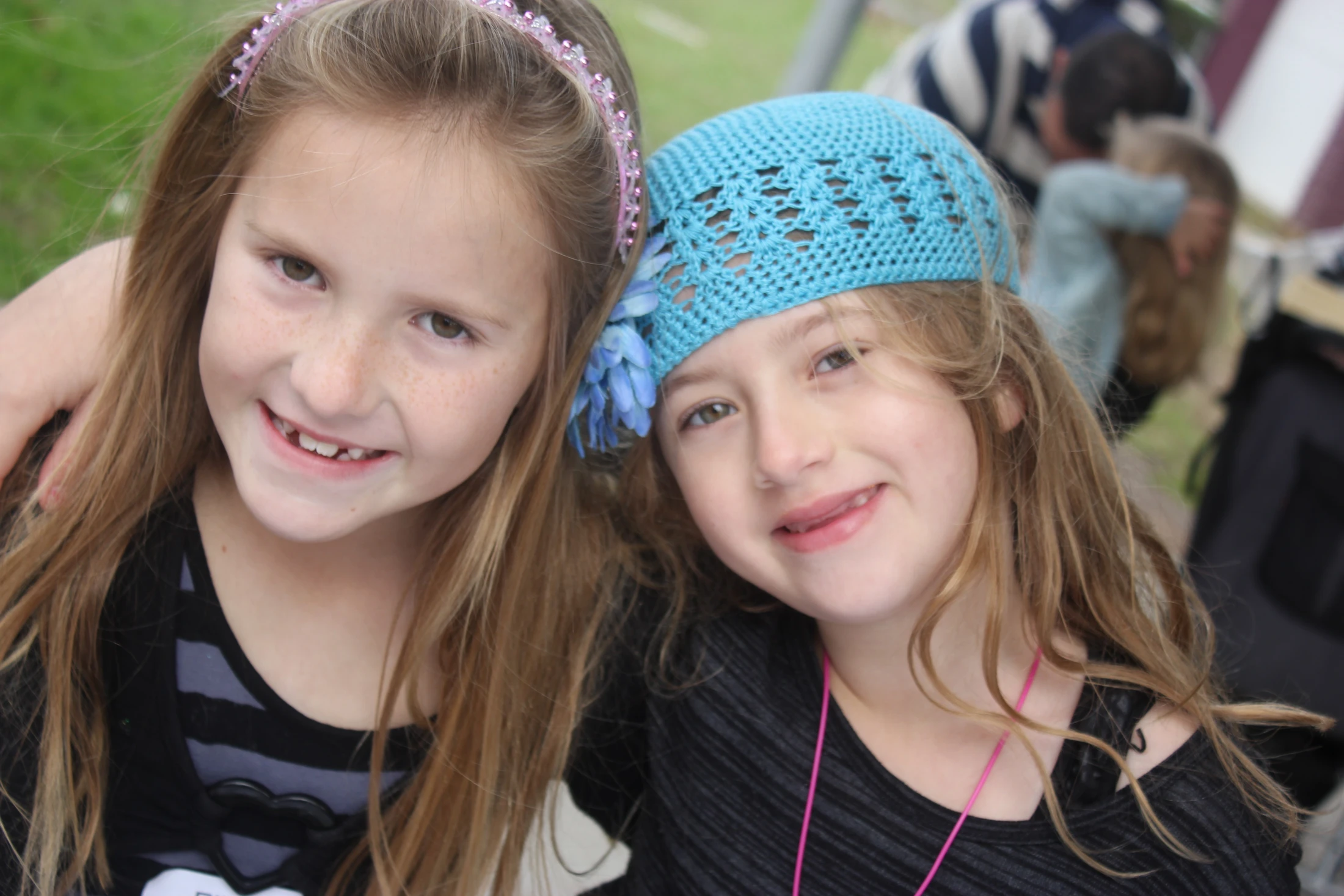 A close-up of two young girls smiling at the camera. The girl on the left has a pink headband, and the girl on the right wears a blue crocheted headband.