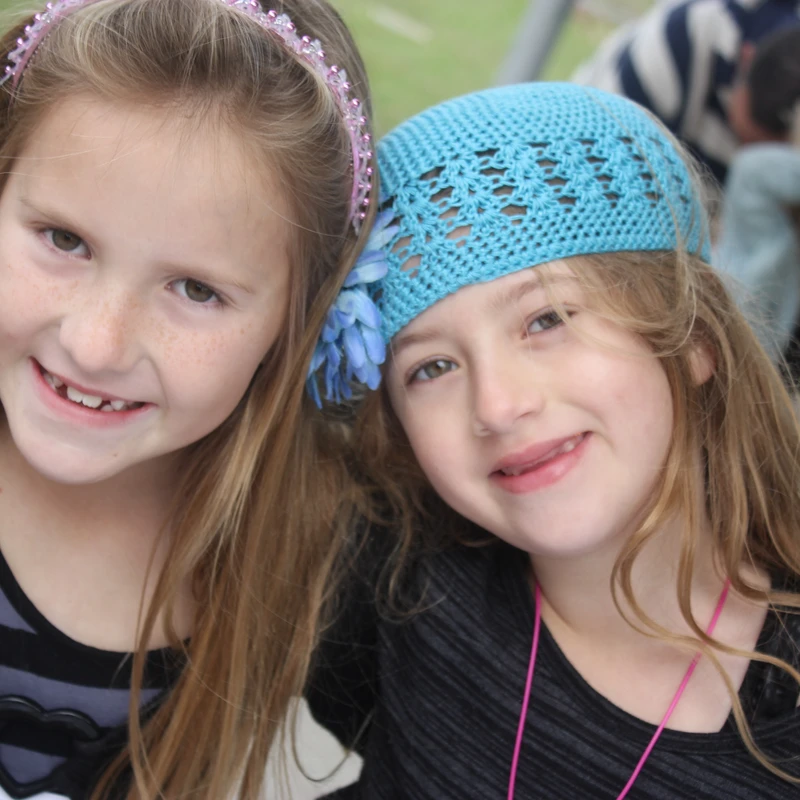 A close-up of two young girls smiling at the camera. The girl on the left has a pink headband, and the girl on the right wears a blue crocheted headband.
