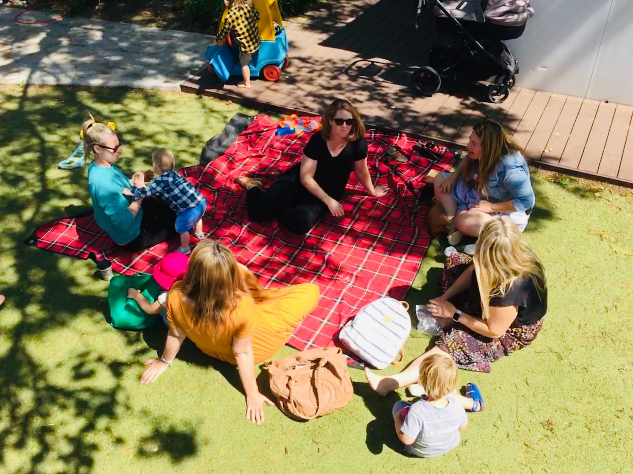 A high-angle view of a group of women and young children sitting on a red checkered blanket on a sunny lawn, enjoying a picnic together under dappled shade.
