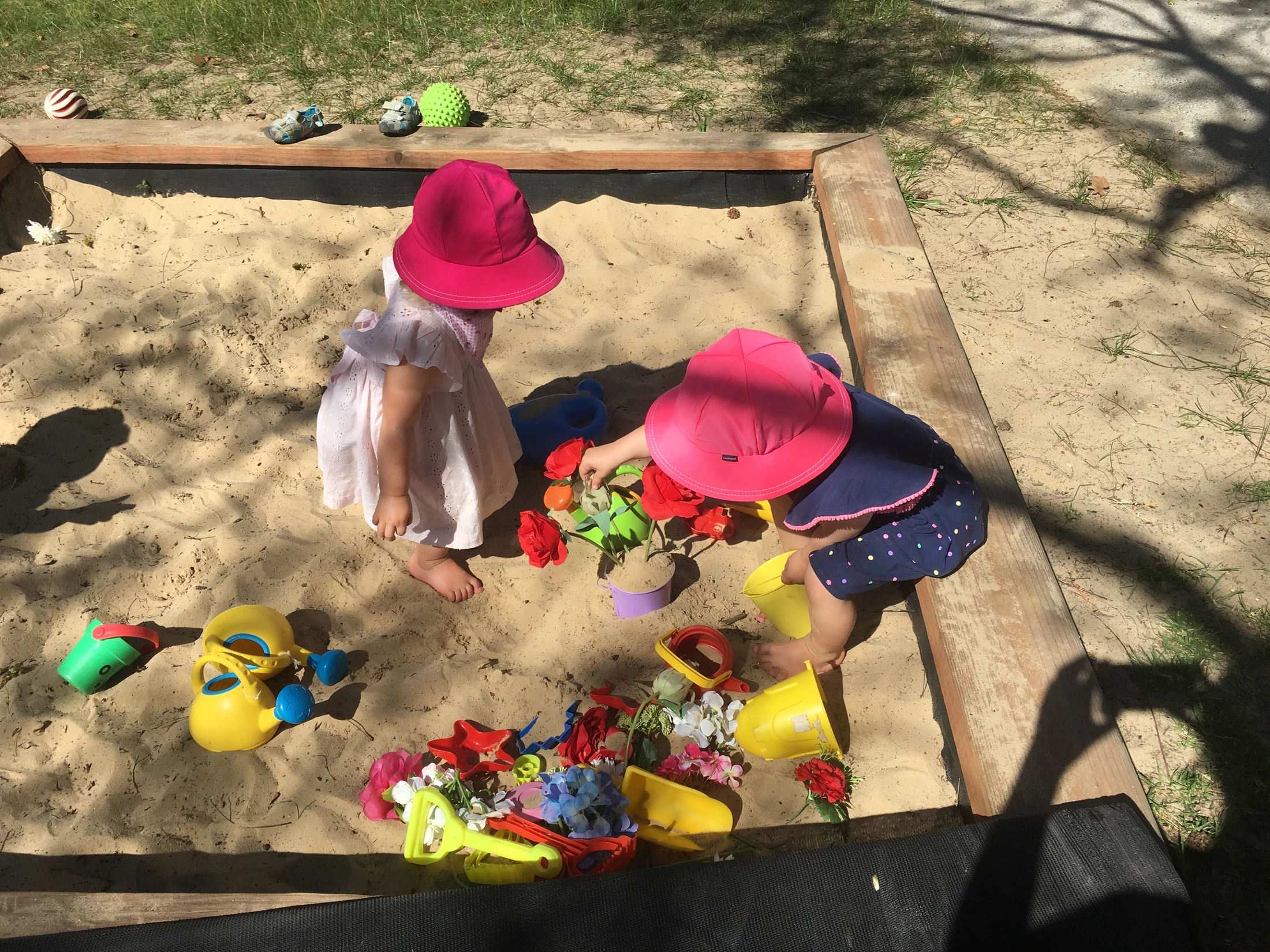 An overhead view of two toddlers in pink sun hats playing with colorful plastic toys and artificial flowers in a large wooden sandbox.
