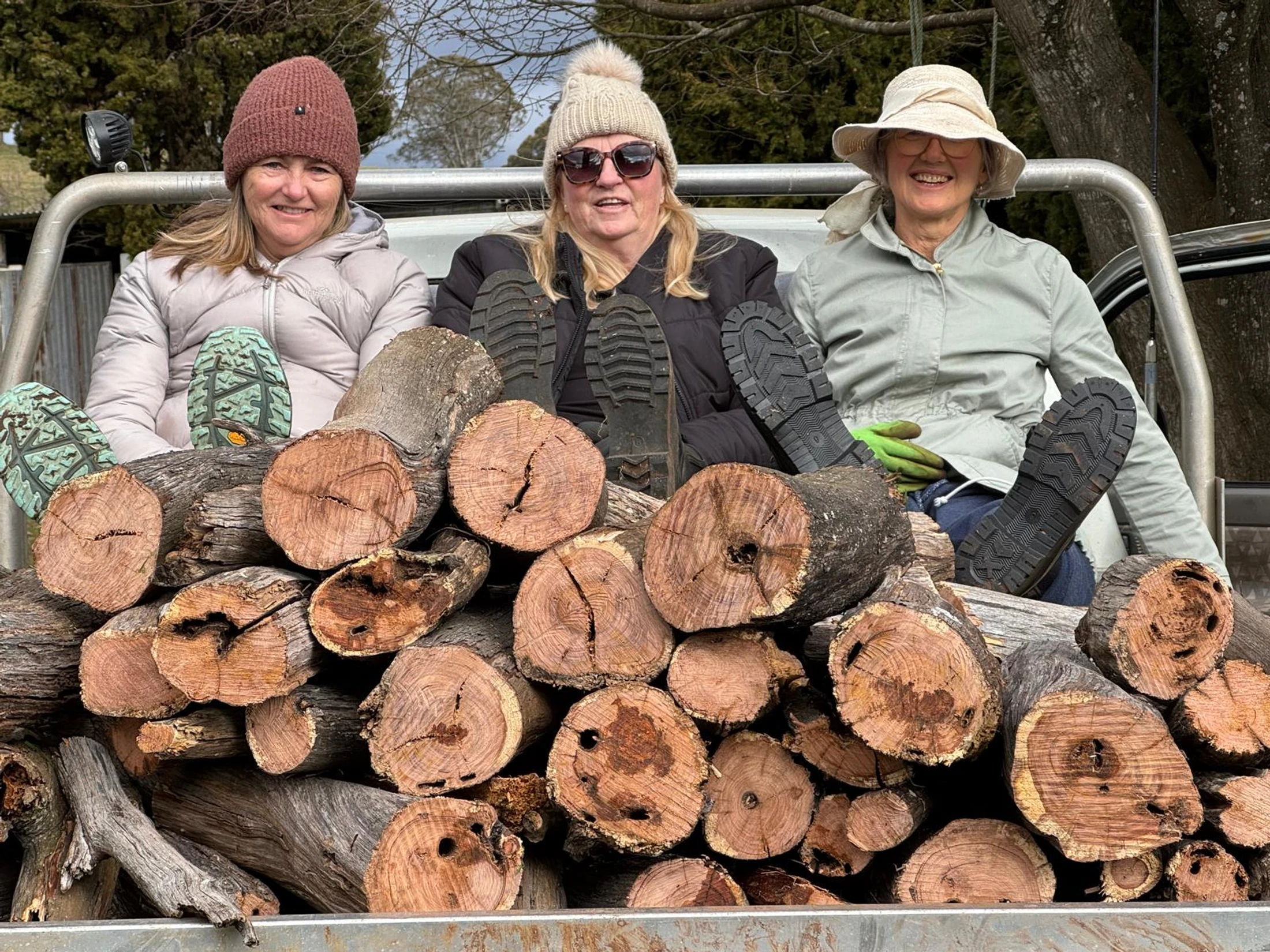 Three smiling women in warm hats and jackets sit in a truck bed, resting their feet on a large pile of freshly cut firewood logs in front of them.