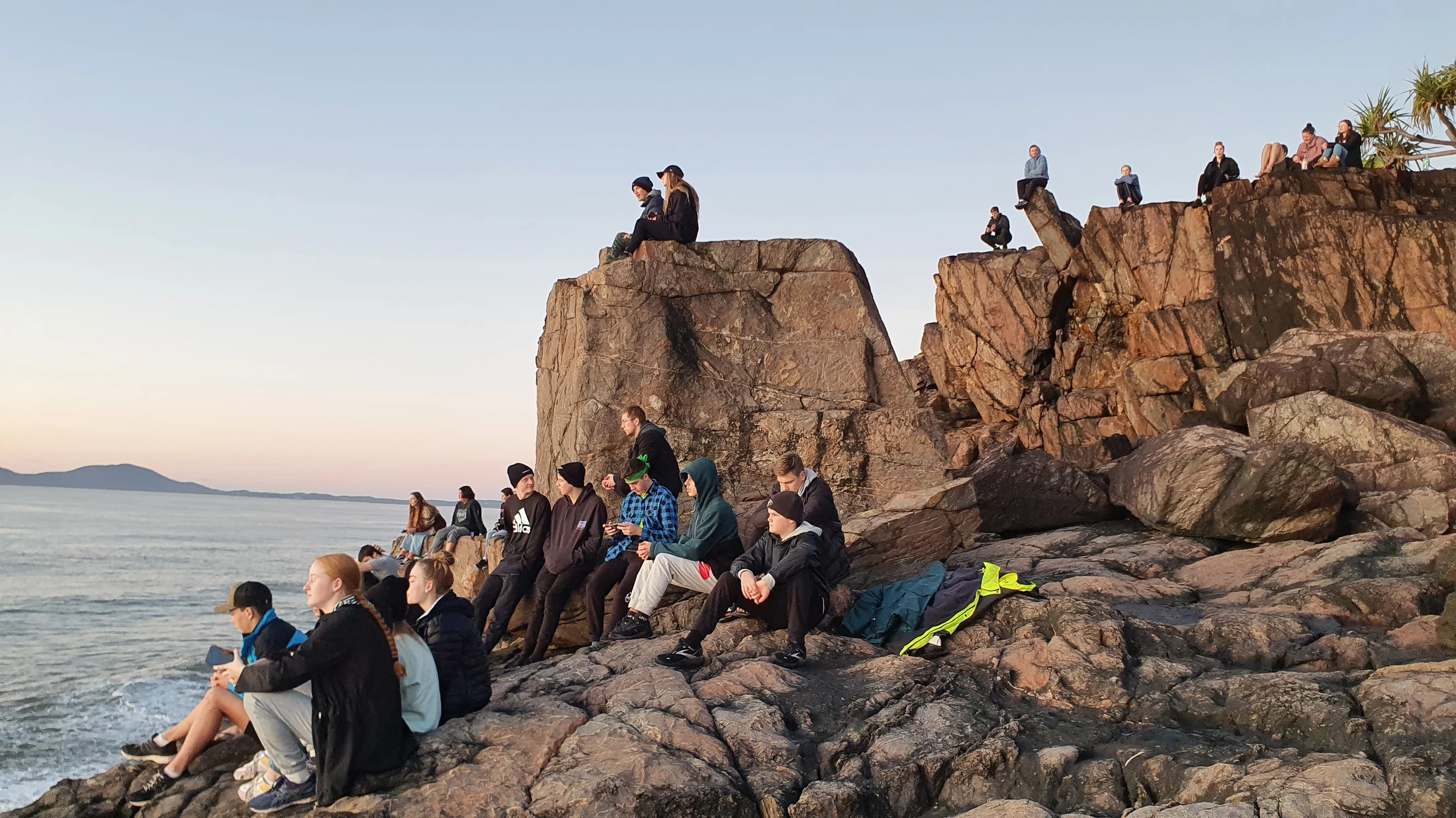 A large group of young people sits scattered across a rocky headland by the ocean, watching the sunset with distant hills on the horizon.