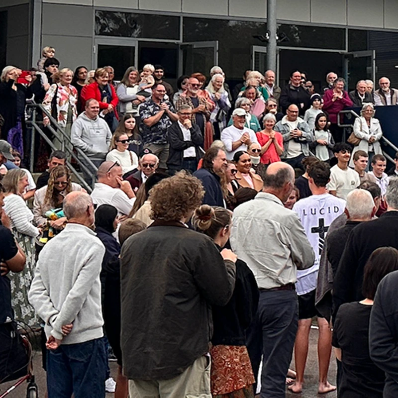A large, diverse crowd of people of all ages gathered outdoors in front of a building. Some are on a raised platform, others are below, all watching something.