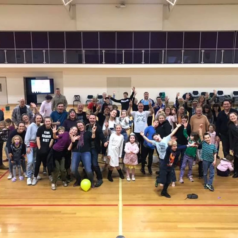 A large group of adults and children posing happily for a photo on the wooden floor of an indoor gymnasium. Many are smiling and waving at the camera.