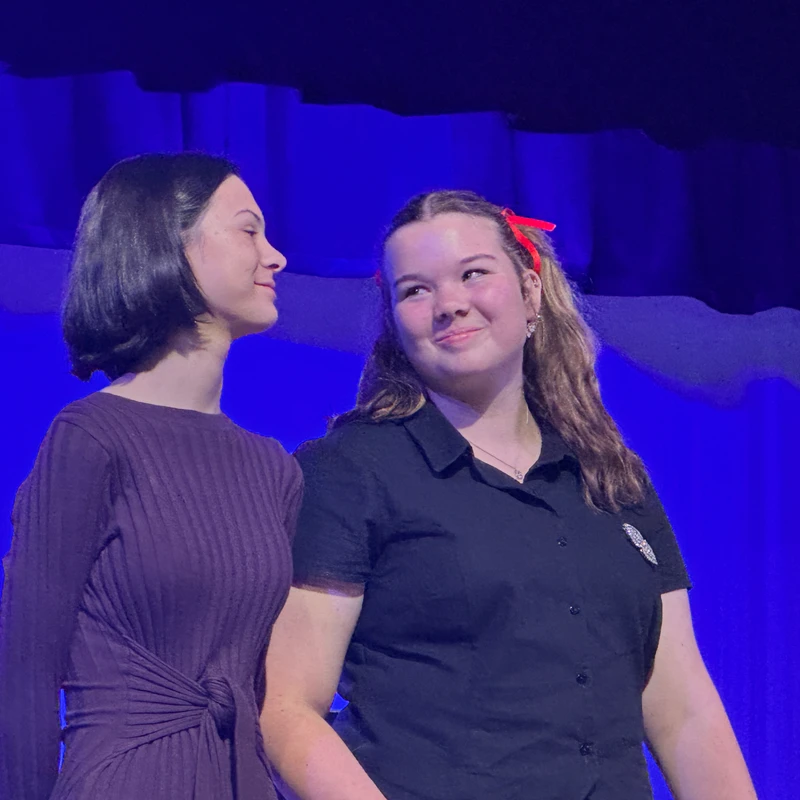 Two young women smile at each other while standing on a stage. The woman on the left has a dark bob, and the woman on the right has a red ribbon in her hair.