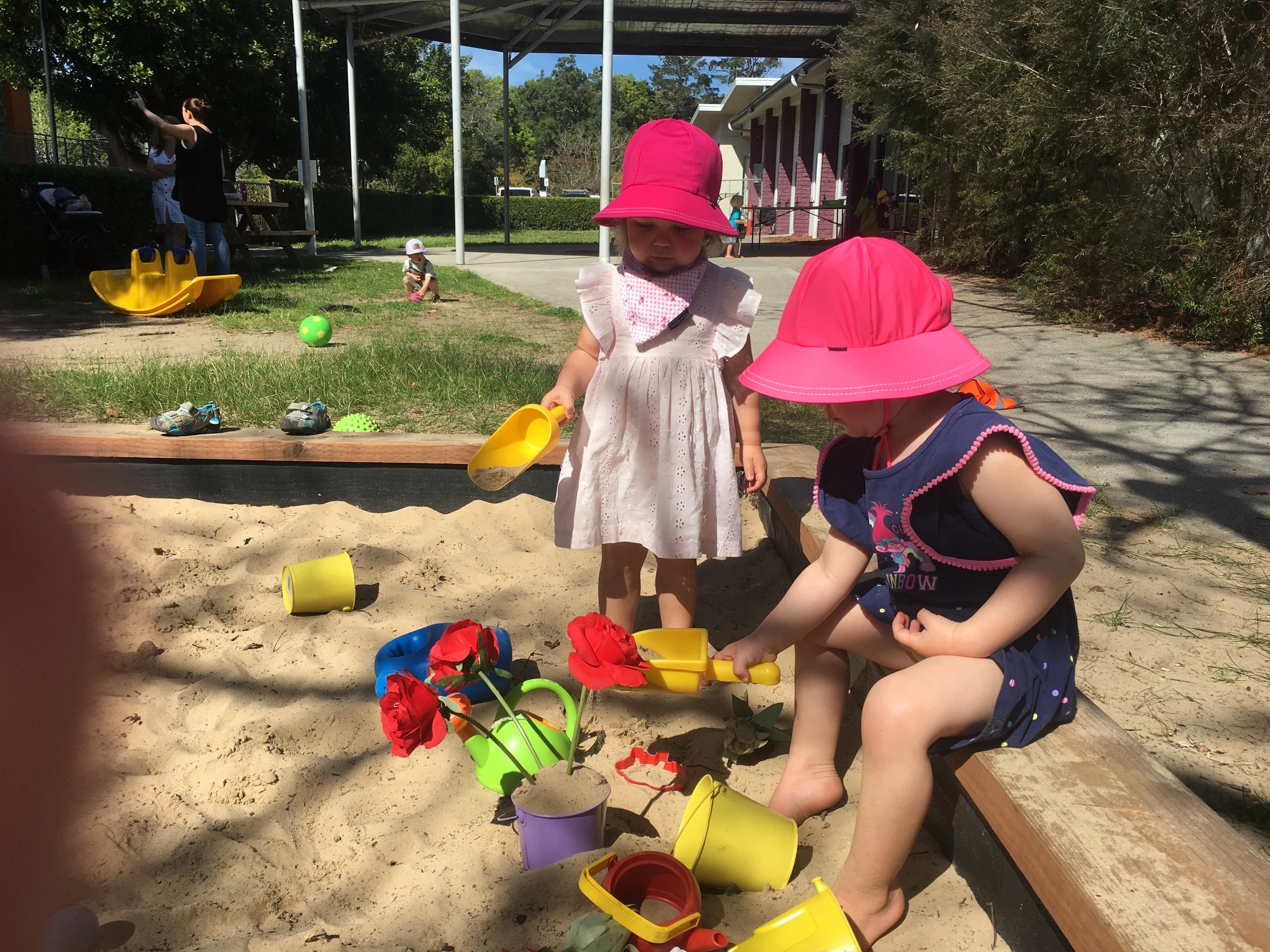 Two young children wearing matching pink sun hats play with colorful plastic toys in a large sandbox on a sunny day at a playground.