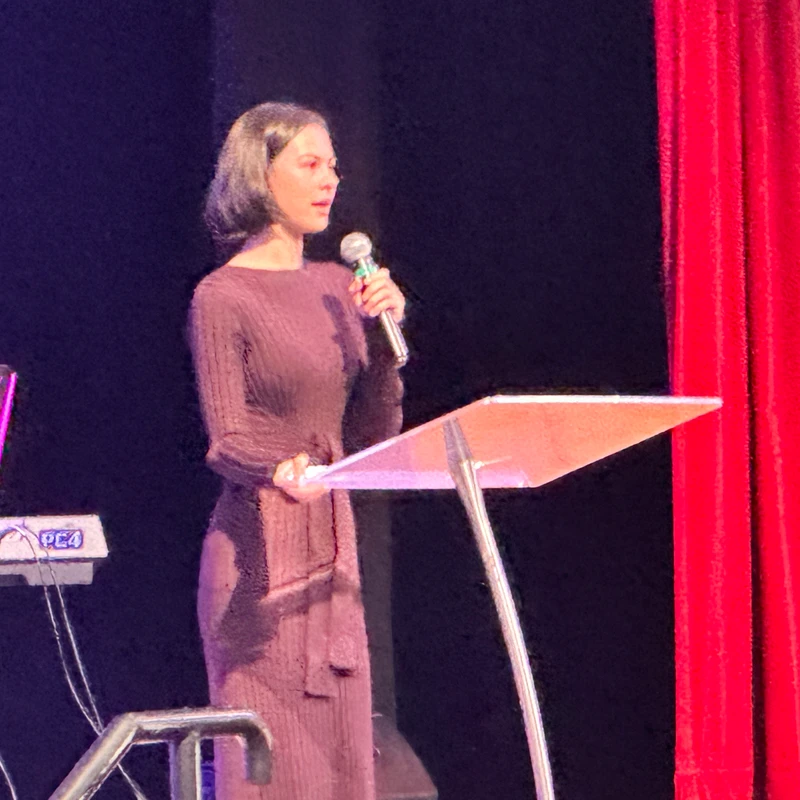 A woman with short dark hair in a long-sleeved purple dress stands at a podium on a stage, holding a microphone and speaking.