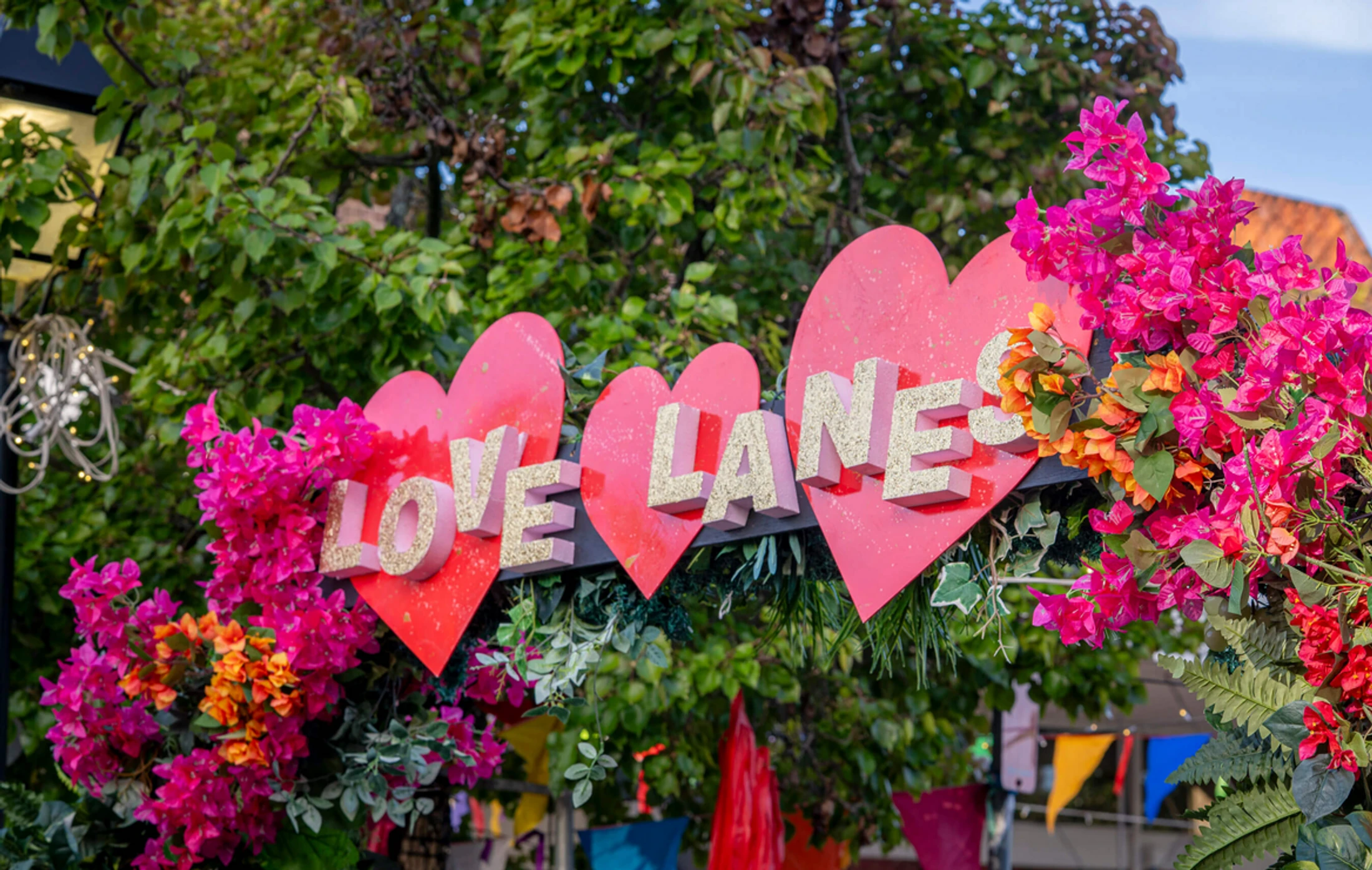 A colorful sign with the words 'Love Lanes' in glittery letters on red hearts, adorned with bright pink and orange flowers against a backdrop of green trees.
