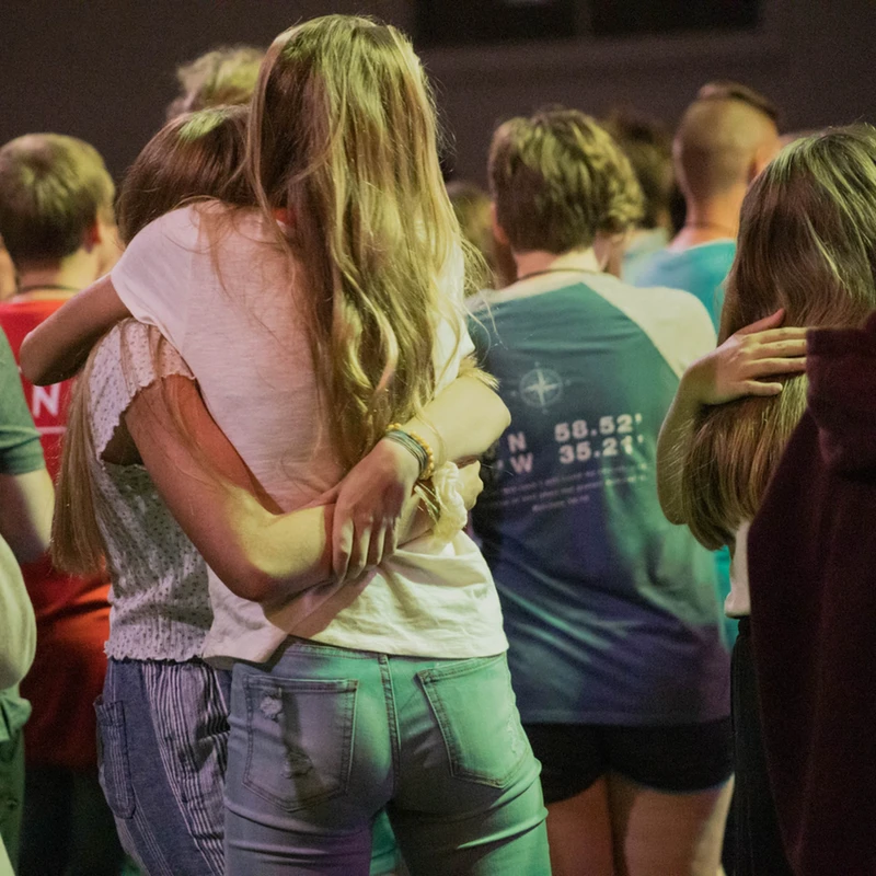A view from behind of two young women with long hair hugging in the middle of a crowd at an event, surrounded by other young people.