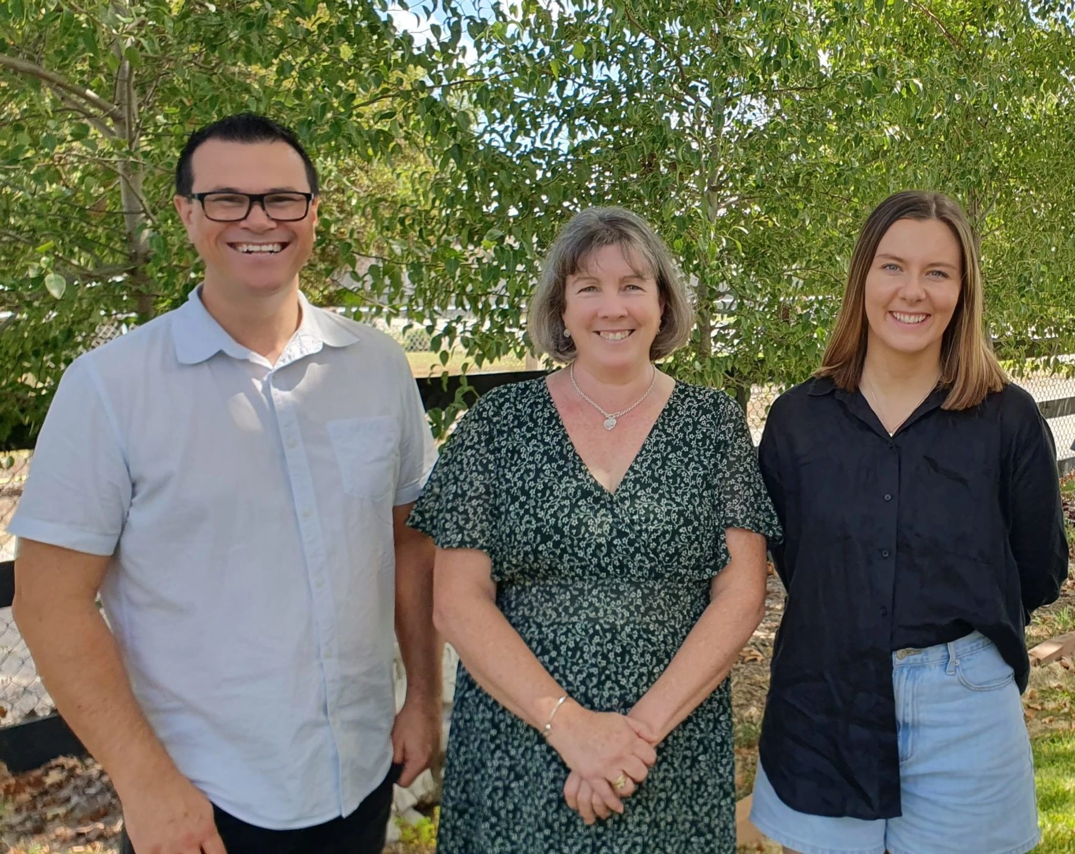 A man and two women stand side by side outdoors in front of a leafy green tree, smiling for the camera on a sunny day.