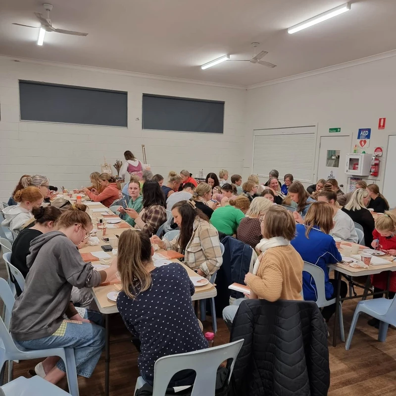 A large group of women and children gathered in a community hall. They are sitting at long tables, focused on an arts and crafts activity together.