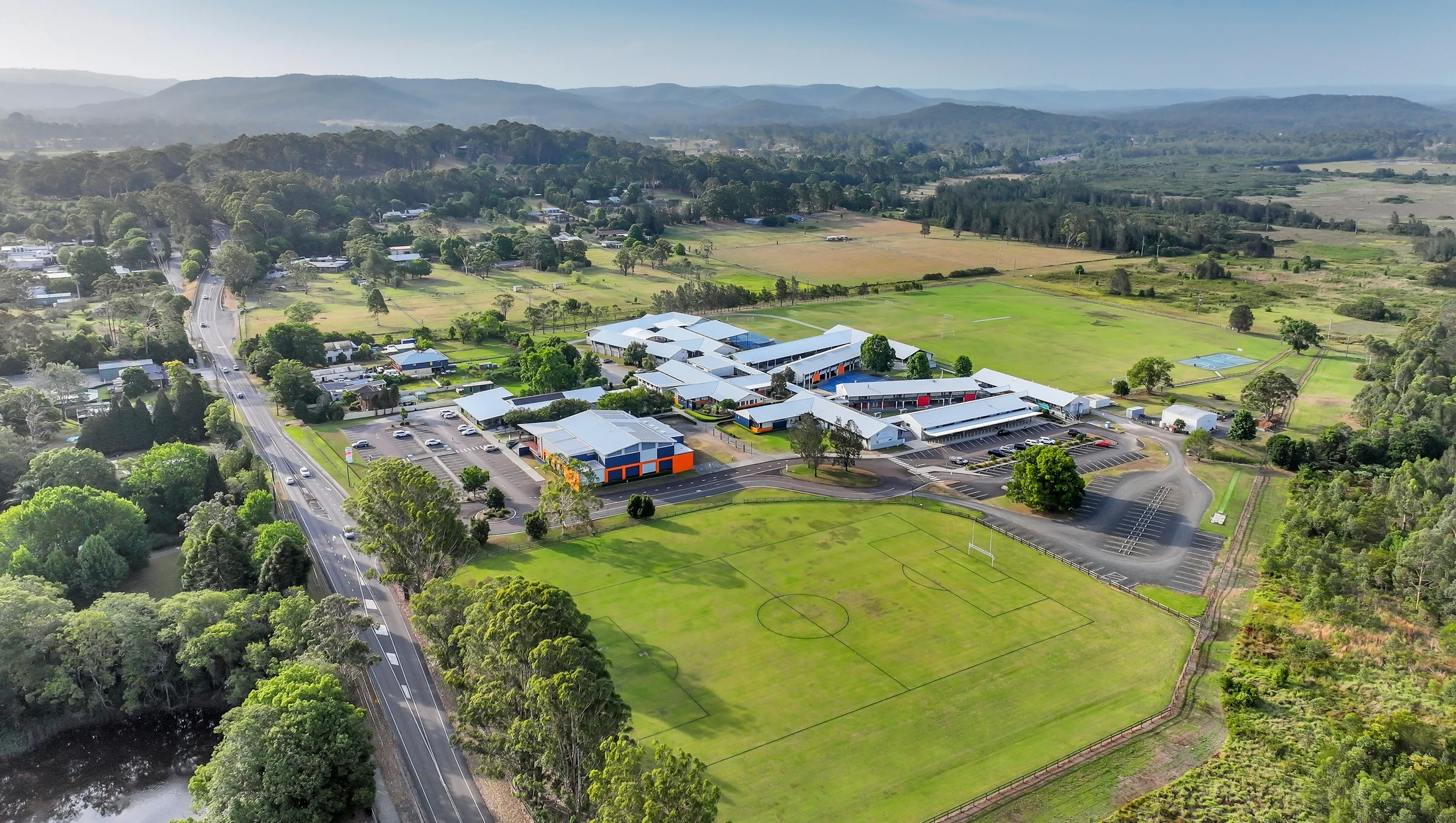 An aerial view shows a sprawling school campus with sports fields, nestled in a green valley with houses, a road, and mountains in the background.