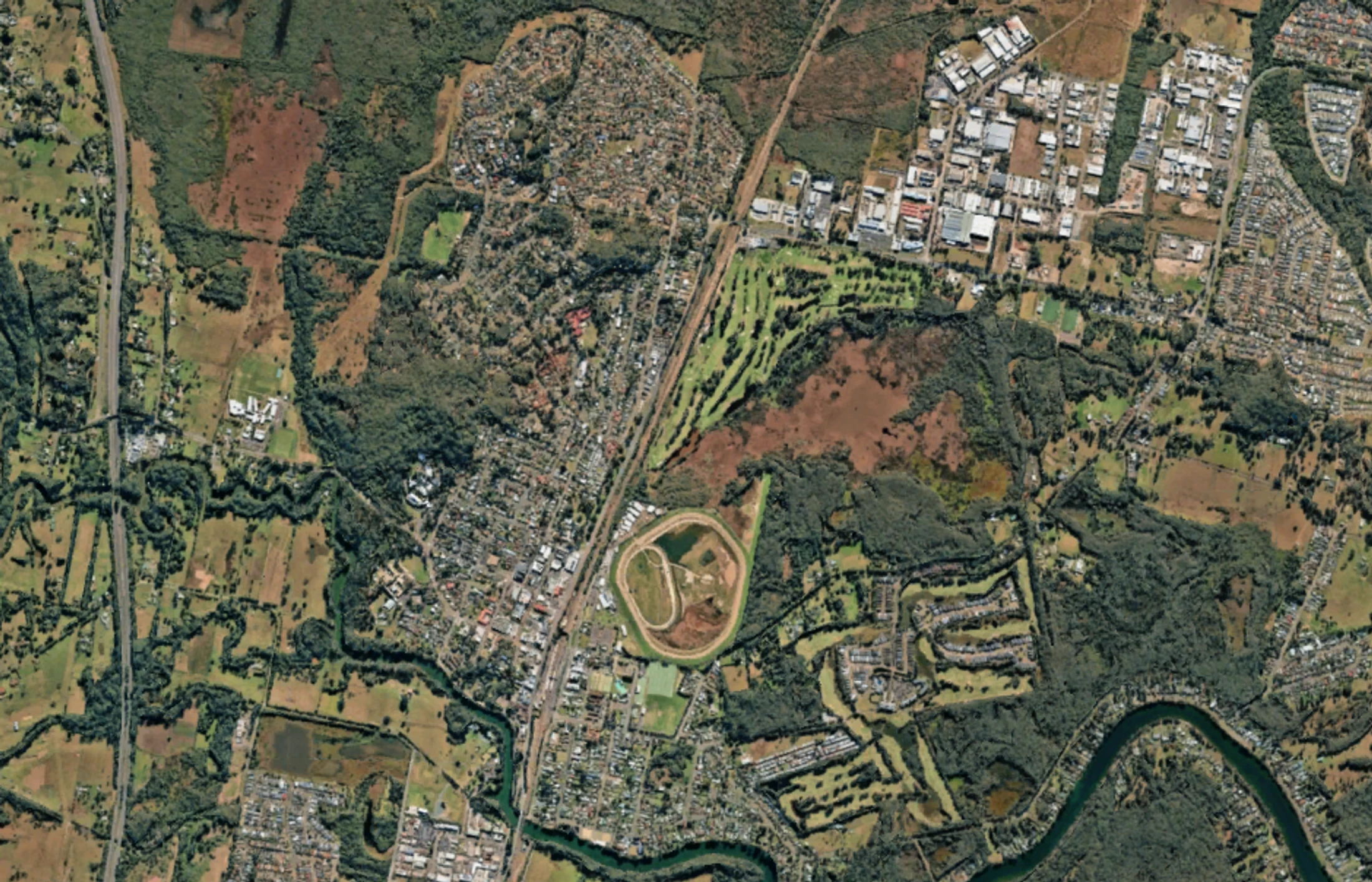 An overhead satellite photo showing a town nestled among green forests. A highway, a railway, a winding river, a golf course, and a racetrack are visible.