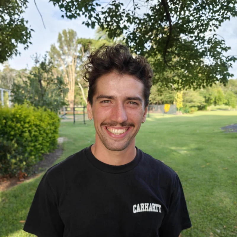 A young man with dark wavy hair and a mustache smiles while standing outside in a park. He wears a black Carhartt t-shirt, and trees are in the background.