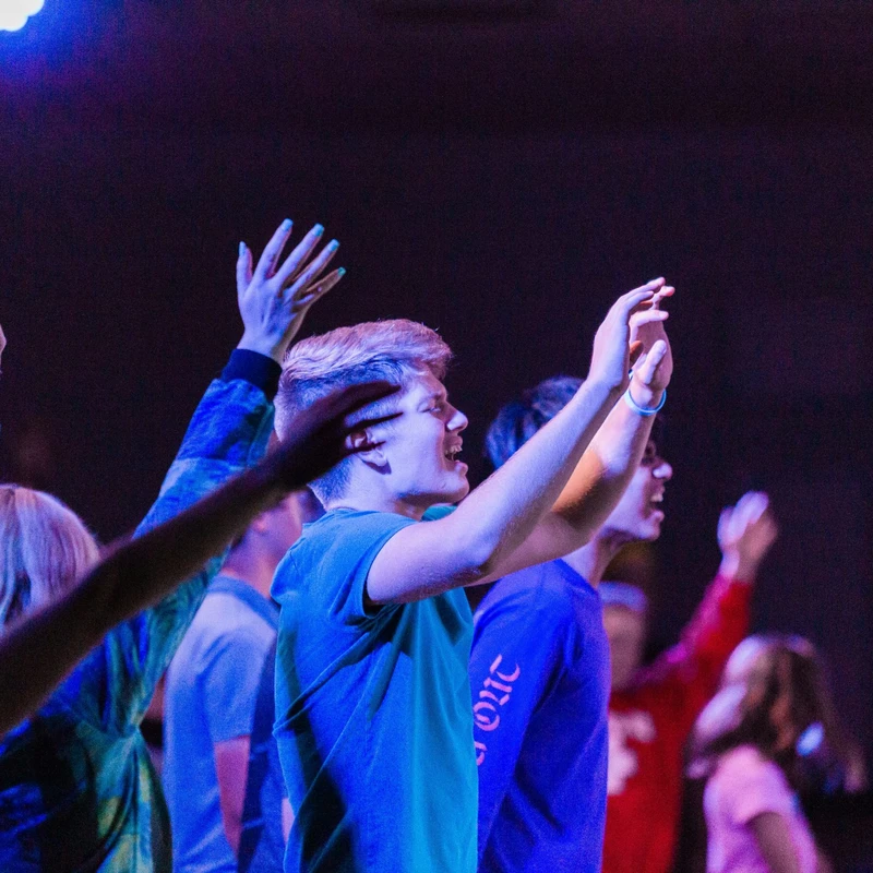 A young man in a green shirt sings with his hands raised in a crowd of people worshiping under blue and purple lights.