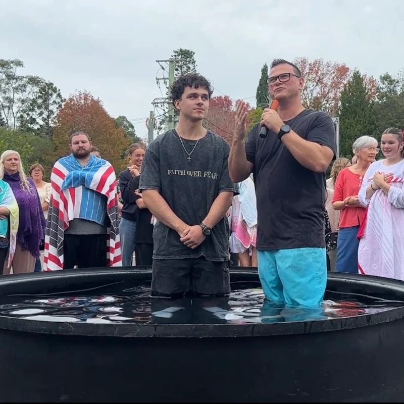 A pastor with a microphone stands in a large tub of water with a young man during an outdoor baptism ceremony, with a crowd of people watching on.