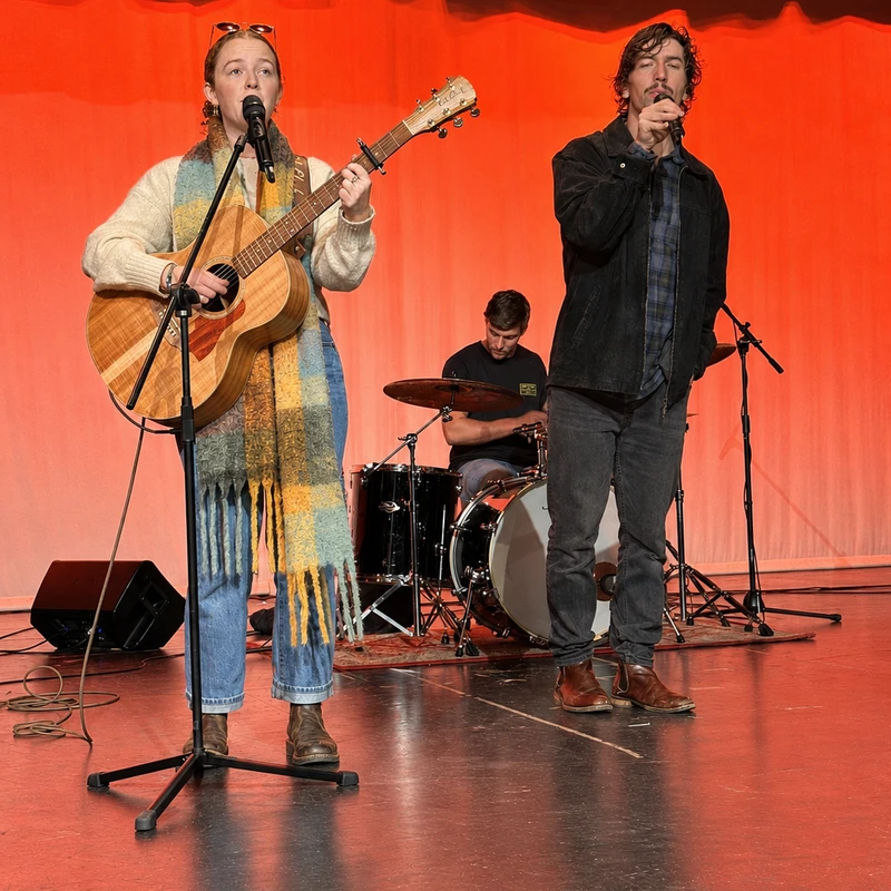A band with a female guitarist and male singer at the front and a drummer in the back performs on a stage lit with red light against a red curtain.
