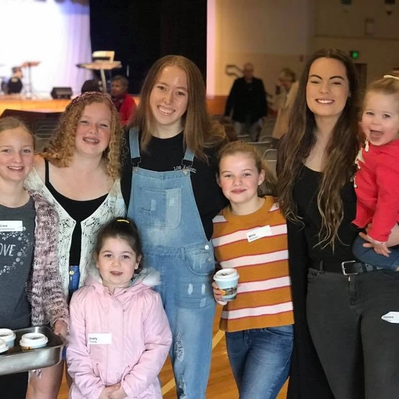 A group of seven girls and young women of different ages smile for a photo in a large hall. One woman holds a happy toddler in a red hoodie.
