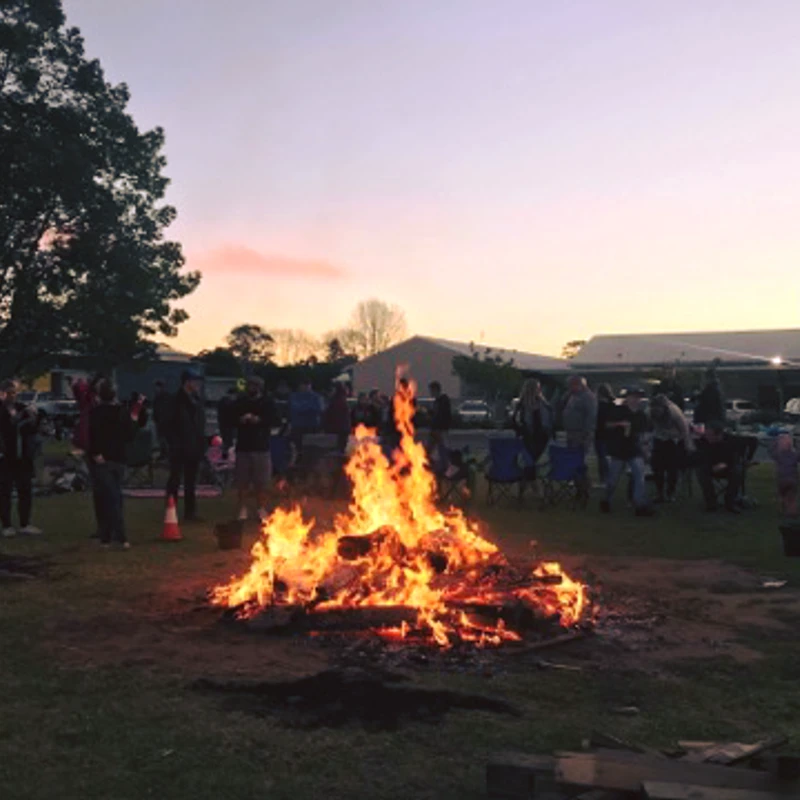 A large group of people gathers on a grassy field at dusk, standing around a roaring bonfire with buildings and trees in the background.