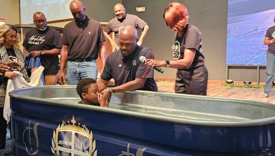 A young Black boy is being baptized by a pastor in a large blue tub on a church stage. Family and congregation members watch and support them.