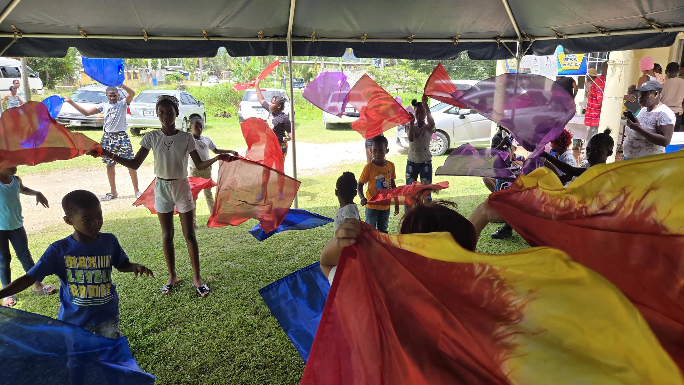 A group of children joyfully dances with colorful red, purple, and blue flags on a grassy lawn under a large black tent during an outdoor community event.