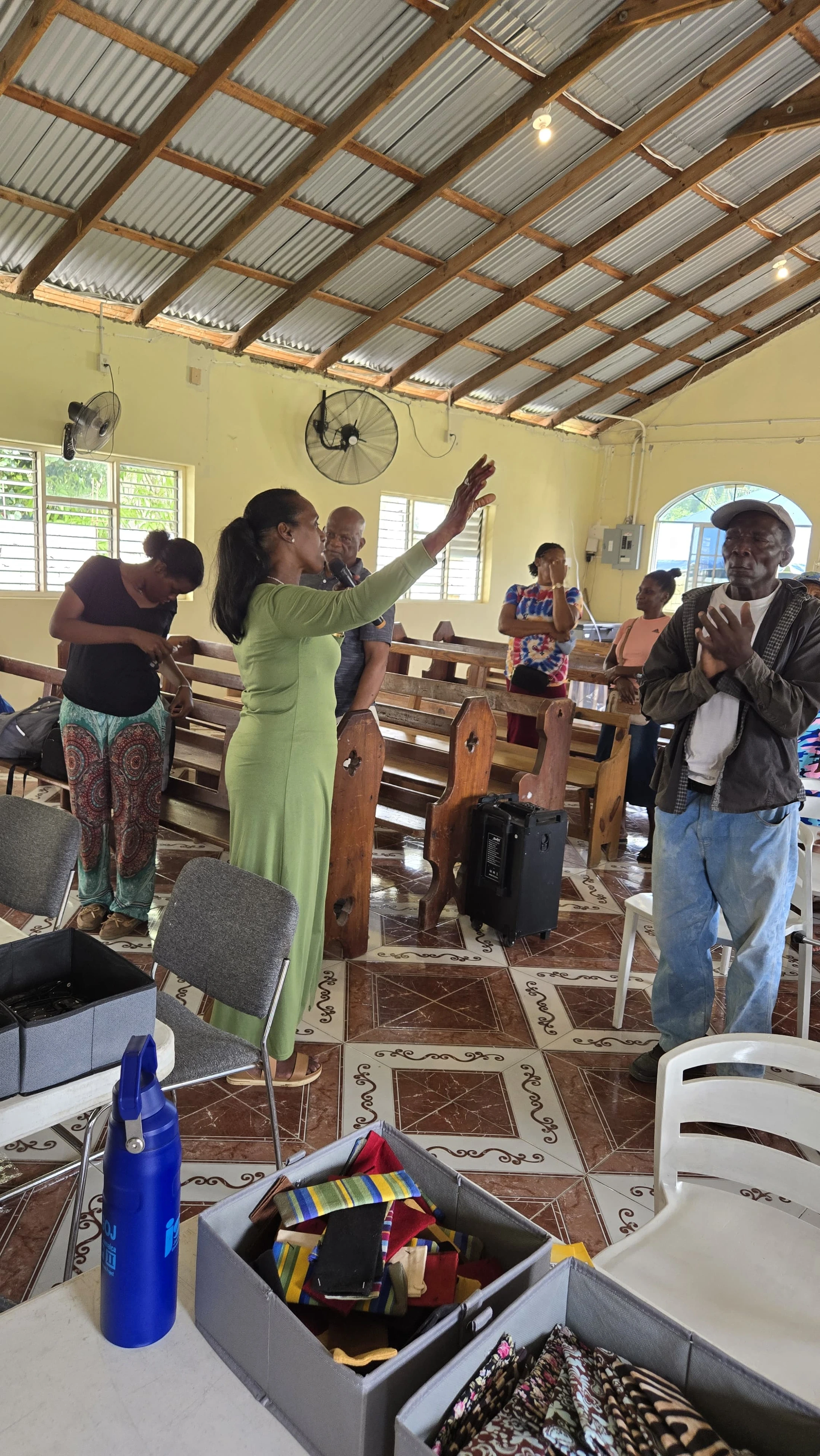 A woman in a green dress leads a diverse group of people in prayer inside a church with wooden pews, tiled floors, and a corrugated metal roof.