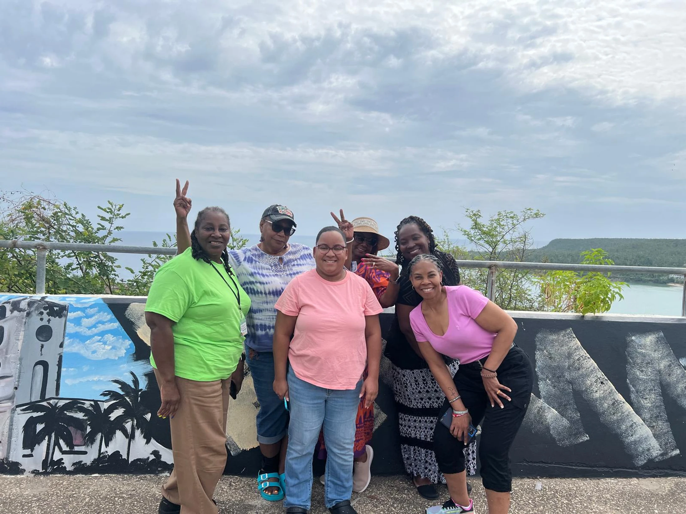 A group of six smiling women pose for a picture in front of a painted wall with a scenic coastal view of the ocean and hills in the background.