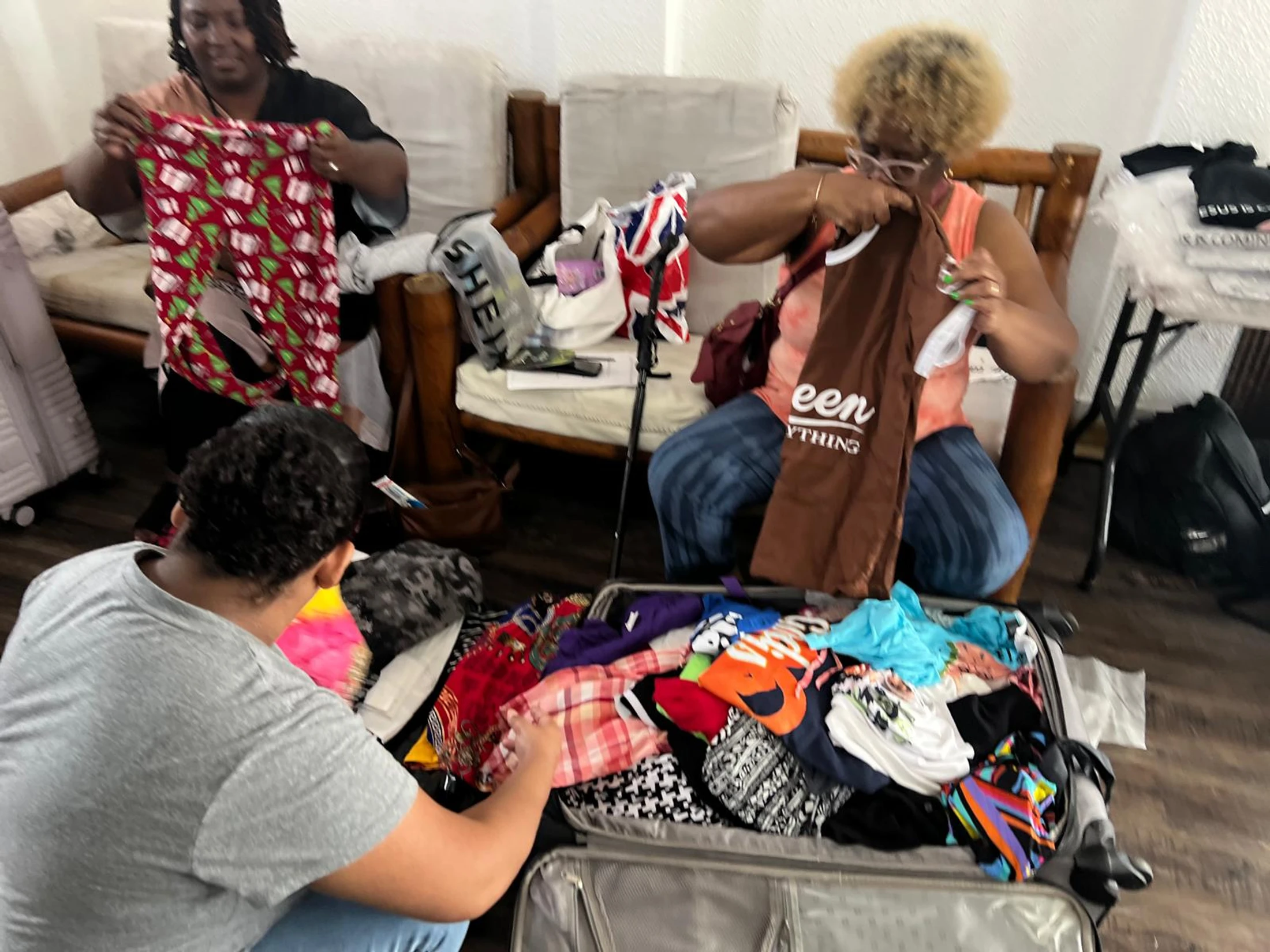 Three women are gathered in a room, sorting through colorful clothes and packing them into an open suitcase on the floor.