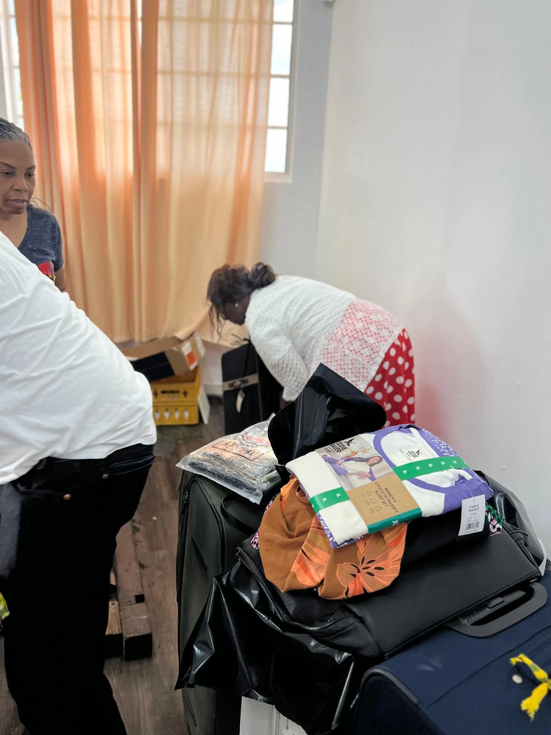 Two women sort through items and pack suitcases in a room. In the foreground, luggage is piled with new packaged clothing and other goods.